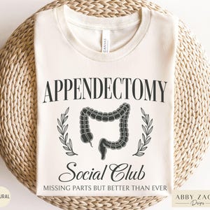 May include: Cream-colored t-shirt with the words "Appendectomy Social Club" in black, featuring a stylized illustration of an appendix and laurel leaves. The text "Missing parts but better than ever" is below the illustration.