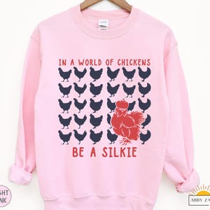 May include: A pink sweatshirt with the text "In a world of chickens be a silkie" in blue and red lettering. The design features a red silkie chicken surrounded by black chickens.