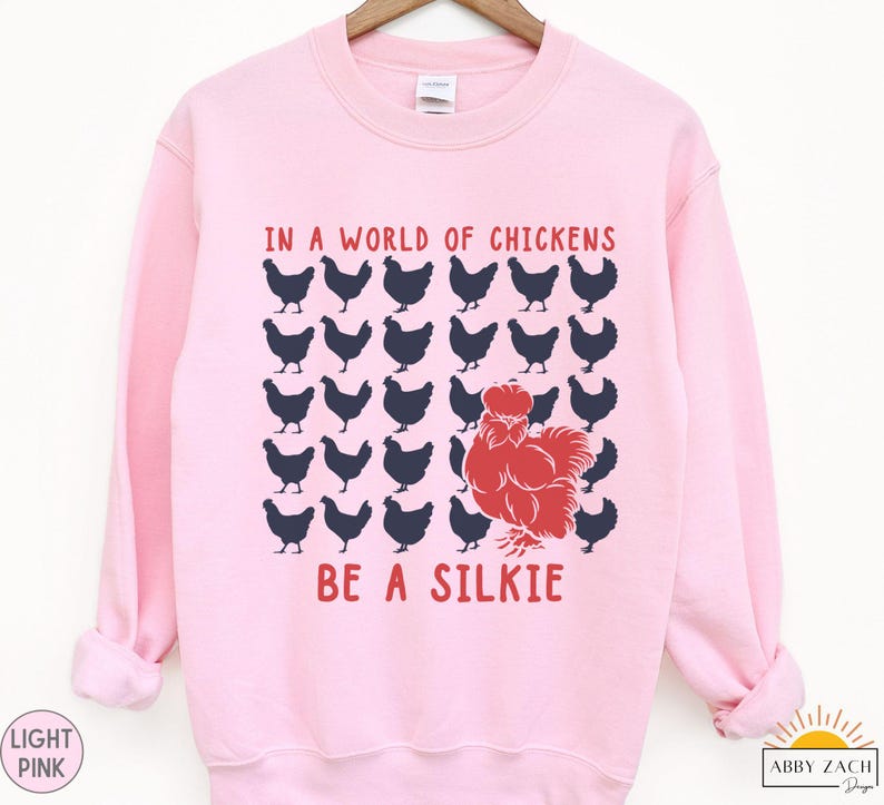 May include: A pink sweatshirt with the text "In a world of chickens be a silkie" in blue and red lettering. The design features a red silkie chicken surrounded by black chickens.