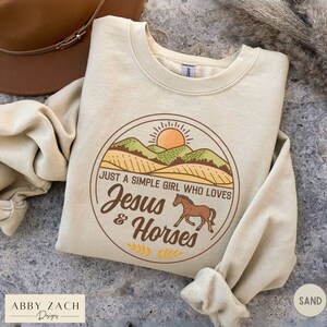 K&ouml;nnte beinhalten: Ein beiger Pullover mit einem Grafikdesign einer Sonne, Bergen und einem Pferd. Die Grafik lautet "Just a simple girl who loves Jesus & Horses".