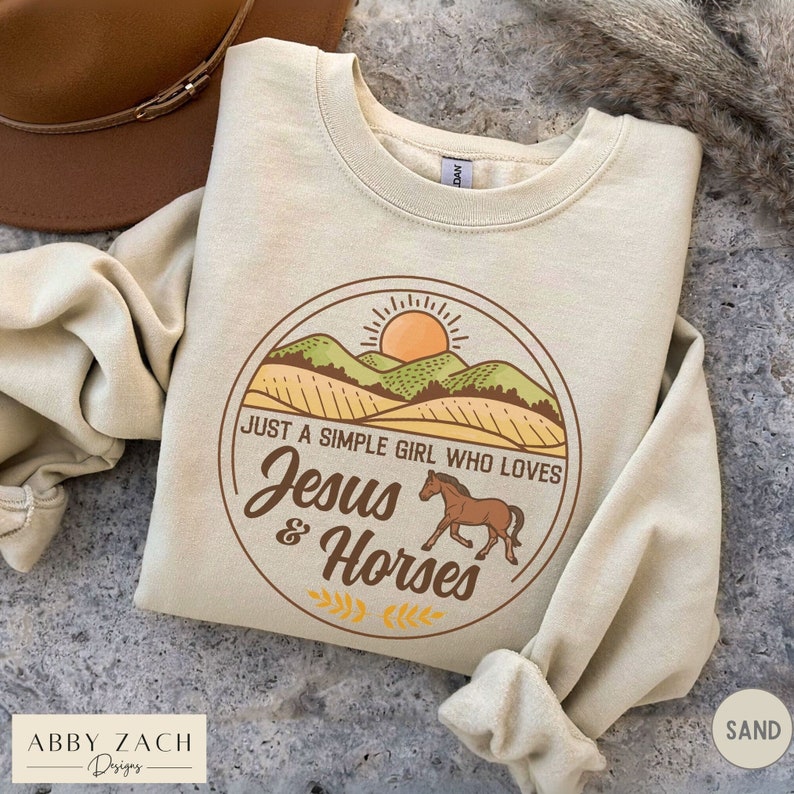 K&ouml;nnte beinhalten: Ein beiger Pullover mit einem Grafikdesign einer Sonne, Bergen und einem Pferd. Die Grafik lautet "Just a simple girl who loves Jesus & Horses".