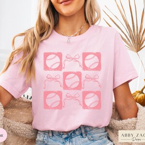 May include: Pink t-shirt with a graphic design of baseballs and bows in a grid pattern. The design is in a distressed style.
