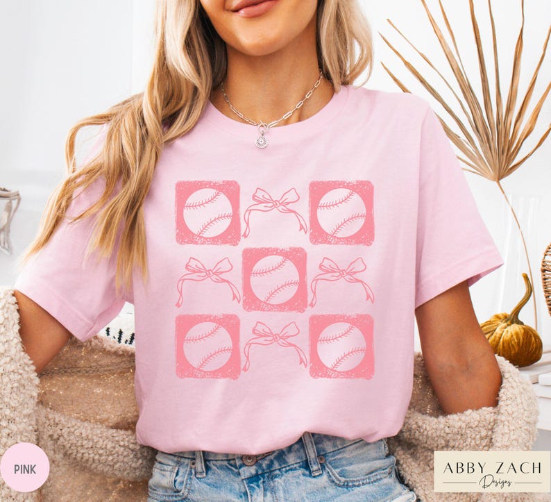 May include: Pink t-shirt with a graphic design of baseballs and bows in a grid pattern. The design is in a distressed style.