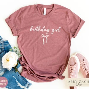May include: Heather mauve t-shirt with the words "birthday girl" in cursive script and a bow graphic. The shirt is paired with blue jeans and pink sneakers. The shirt is a casual wear item.