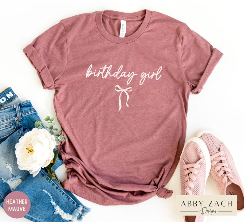 May include: Heather mauve t-shirt with the words "birthday girl" in cursive script and a bow graphic. The shirt is paired with blue jeans and pink sneakers. The shirt is a casual wear item.