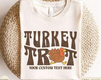 May include: A cream-colored t-shirt with the words "TURKEY TROT" in brown, retro-style lettering. A cartoon turkey in brown, red, and yellow is running below the text. The phrase "YOUR CUSTOM TEXT HERE" is below the turkey.
