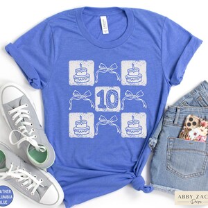 Peut inclure: Un t-shirt bleu avec un motif blanc repr&eacute;sentant un g&acirc;teau d'anniversaire et le chiffre 10. Le motif est r&eacute;p&eacute;t&eacute; en grille.