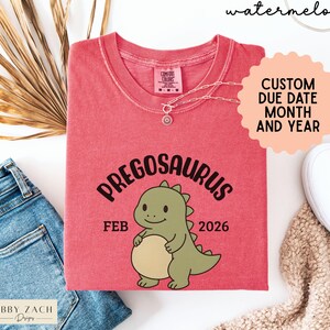 K&ouml;nnte beinhalten: Ein rotes T-Shirt mit dem Wort "PREGOSAURUS" &uuml;ber einer Cartoon-Dinosaurier-Illustration. Das Shirt zeigt auch den Monat "FEB" und das Jahr "2026". Das Shirt hat einen Rundhalsausschnitt und kurze &Auml;rmel. Das Wort "watermelon" steht oben.