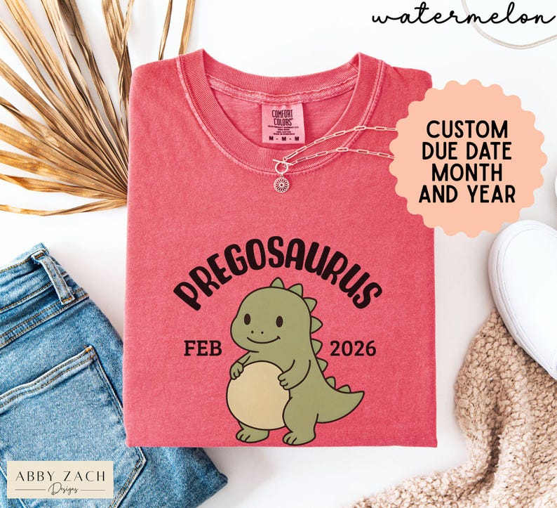 K&ouml;nnte beinhalten: Ein rotes T-Shirt mit dem Wort "PREGOSAURUS" &uuml;ber einer Cartoon-Dinosaurier-Illustration. Das Shirt zeigt auch den Monat "FEB" und das Jahr "2026". Das Shirt hat einen Rundhalsausschnitt und kurze &Auml;rmel. Das Wort "watermelon" steht oben.