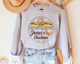 Könnte beinhalten: Grauer Pullover mit einem Grafikdruck eines Sonnenuntergangs über einem Feld mit dem Text "Just a simple girl who loves Jesus & Chickens".