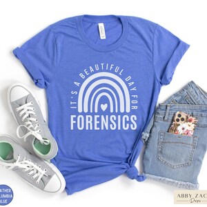May include: A blue t-shirt with a white rainbow and heart design. The text "It's a beautiful day for forensics" is printed on the shirt.
