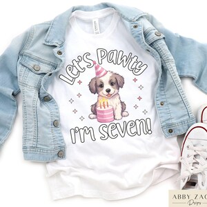 May include: White t-shirt with a cute cartoon dog wearing a party hat and holding a pink cake. The text "Let's Pawty I'm Seven!" is printed on the shirt.