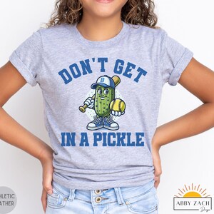 Puede incluir: Una camiseta gris para j&oacute;venes con un pepinillo de dibujos animados que lleva una gorra de b&eacute;isbol y sostiene un bate y una pelota de b&eacute;isbol. El texto "Don't Get In A Pickle" est&aacute; impreso en la camiseta.