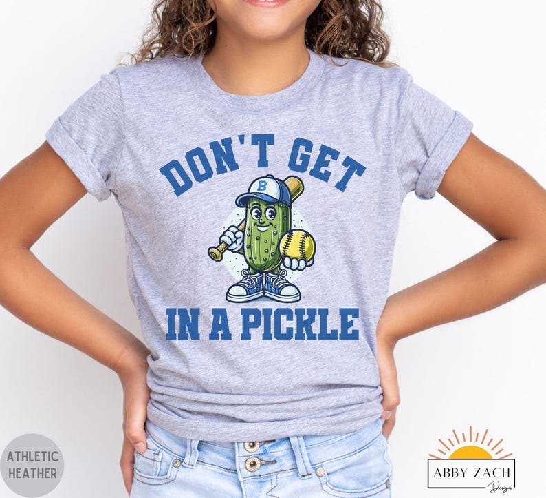 Puede incluir: Una camiseta gris para j&oacute;venes con un pepinillo de dibujos animados que lleva una gorra de b&eacute;isbol y sostiene un bate y una pelota de b&eacute;isbol. El texto "Don't Get In A Pickle" est&aacute; impreso en la camiseta.