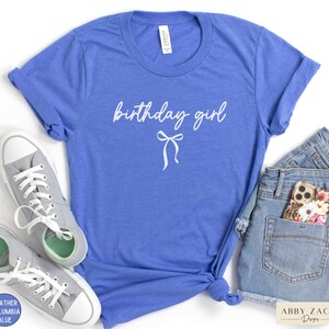 May include: A blue t-shirt with the words "birthday girl" in white script and a bow graphic. The shirt is paired with gray sneakers and denim shorts. The shirt is made of a soft material.