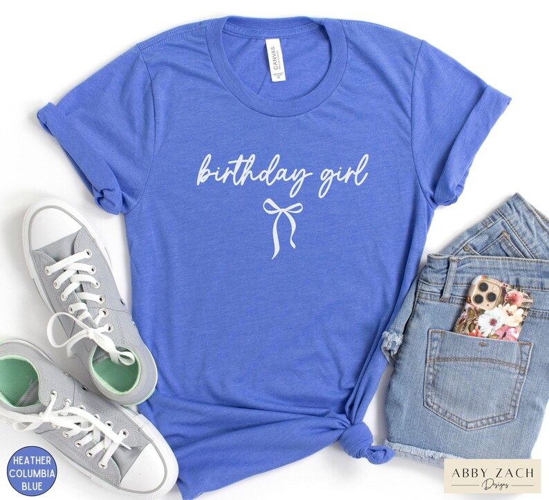 May include: A blue t-shirt with the words "birthday girl" in white script and a bow graphic. The shirt is paired with gray sneakers and denim shorts. The shirt is made of a soft material.