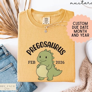 K&ouml;nnte beinhalten: Senffarbenes T-Shirt mit dem Wort "PREGOSAURUS" und einer Cartoon-Dinosaurier-Grafik. Das Shirt zeigt auch den Monat "FEB" und das Jahr "2026". Das Shirt ist von der Marke "COMFORT COLORS".