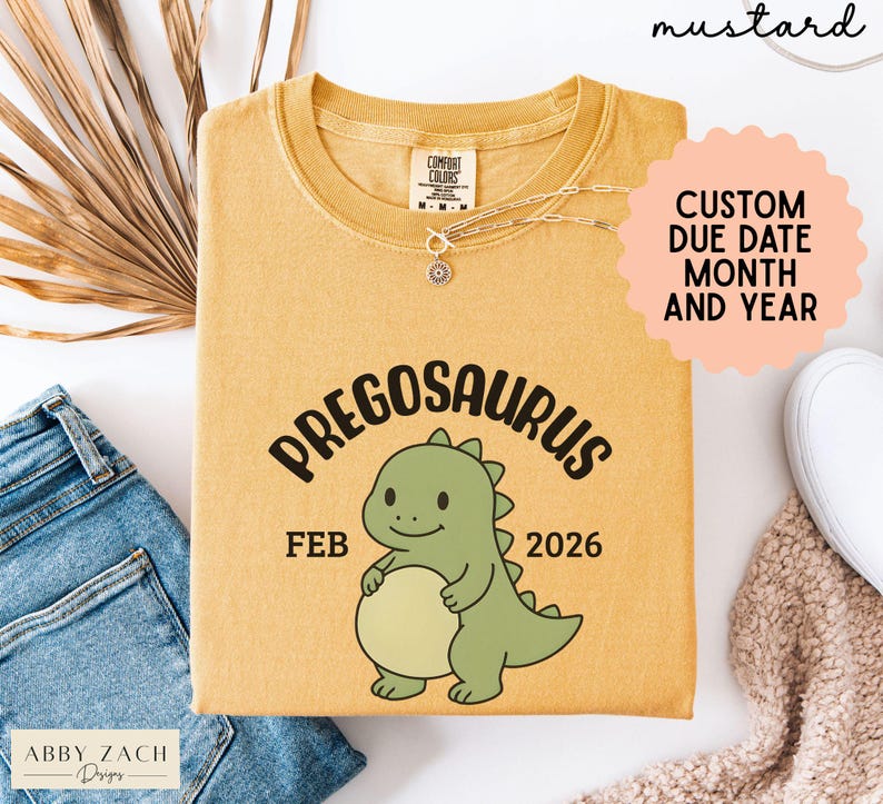K&ouml;nnte beinhalten: Senffarbenes T-Shirt mit dem Wort "PREGOSAURUS" und einer Cartoon-Dinosaurier-Grafik. Das Shirt zeigt auch den Monat "FEB" und das Jahr "2026". Das Shirt ist von der Marke "COMFORT COLORS".