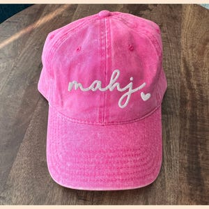 Puede incluir: Gorra de b&eacute;isbol rosa brillante con visera curva. La gorra presenta la palabra "mahj" bordada en cursiva, con un peque&ntilde;o s&iacute;mbolo de coraz&oacute;n. El sombrero est&aacute; hecho de una tela texturizada.