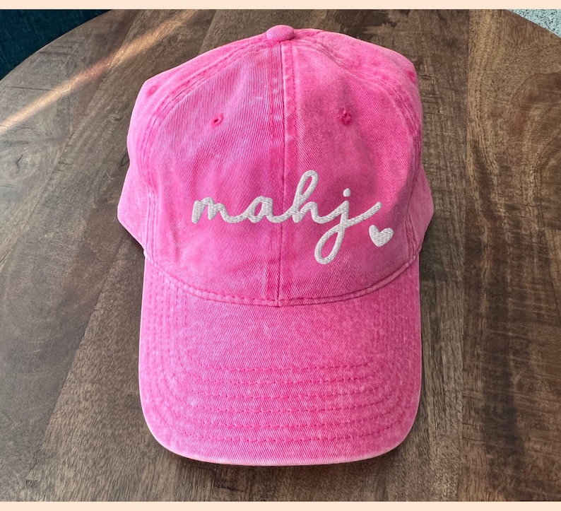 Puede incluir: Gorra de b&eacute;isbol rosa brillante con visera curva. La gorra presenta la palabra "mahj" bordada en cursiva, con un peque&ntilde;o s&iacute;mbolo de coraz&oacute;n. El sombrero est&aacute; hecho de una tela texturizada.