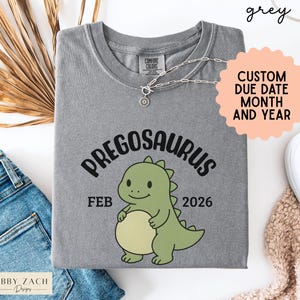 K&ouml;nnte beinhalten: Graues T-Shirt mit dem Wort "PREGOSAURUS" und einer Cartoon-Dinosaurier-Grafik. Das Shirt zeigt auch "FEB 2026" und "CUSTOM DUE DATE MONTH AND YEAR". Marke: Abby Zach Designs.