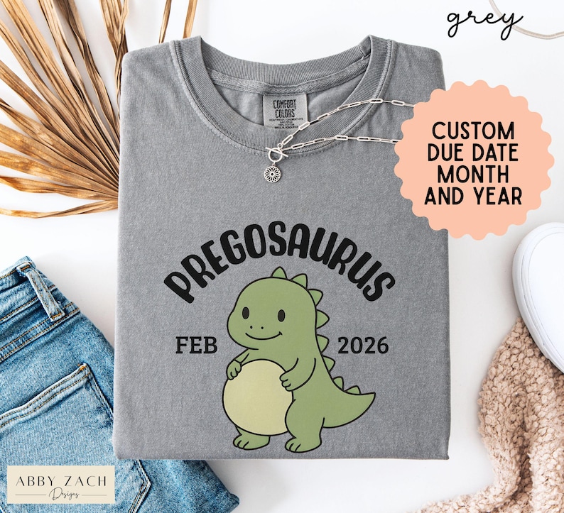 K&ouml;nnte beinhalten: Graues T-Shirt mit dem Wort "PREGOSAURUS" und einer Cartoon-Dinosaurier-Grafik. Das Shirt zeigt auch "FEB 2026" und "CUSTOM DUE DATE MONTH AND YEAR". Marke: Abby Zach Designs.