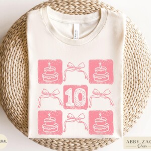 Peut inclure: Un t-shirt blanc avec un motif rose repr&eacute;sentant le chiffre 10 entour&eacute; d'illustrations de g&acirc;teaux roses et de n&oelig;uds. Le texte "Abby Zach Designs" est dans le coin inf&eacute;rieur droit.