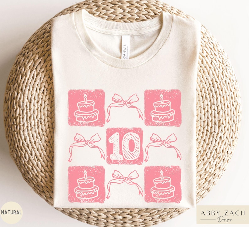 Peut inclure: Un t-shirt blanc avec un motif rose repr&eacute;sentant le chiffre 10 entour&eacute; d'illustrations de g&acirc;teaux roses et de n&oelig;uds. Le texte "Abby Zach Designs" est dans le coin inf&eacute;rieur droit.