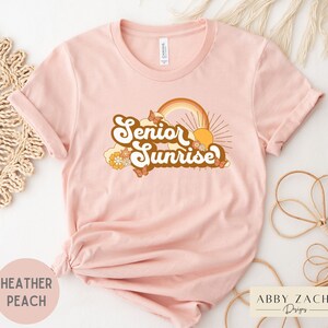 Puede incluir: Una camiseta rosa con el texto "Senior Sunrise" en una fuente retro con un arco&iacute;ris, un sol y mariposas. La camiseta est&aacute; hecha de un material suave y c&oacute;modo.