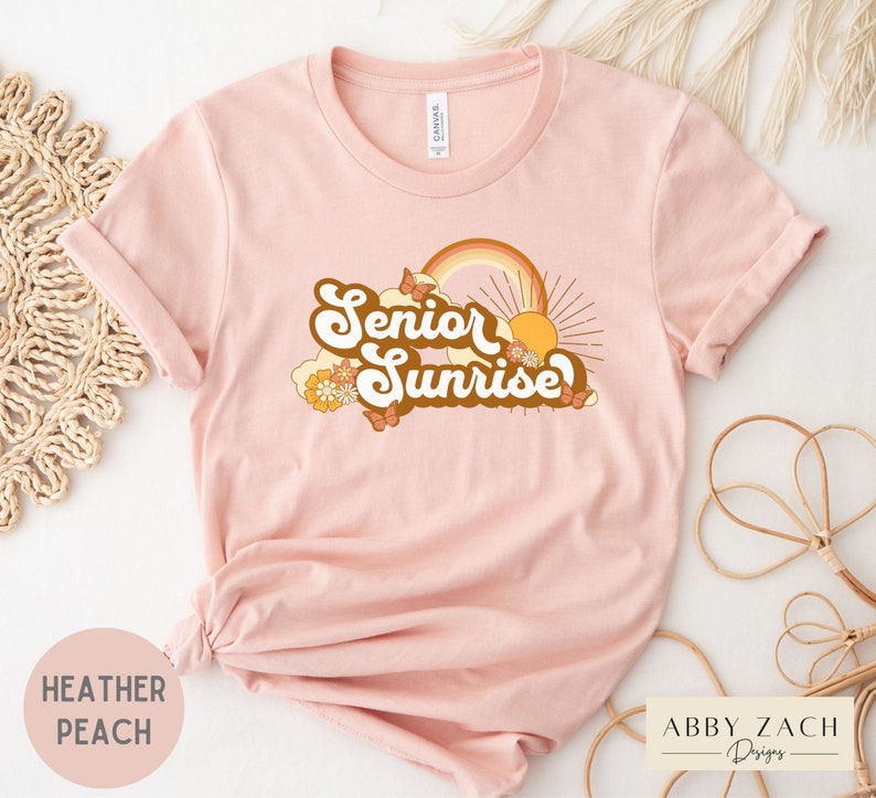 Puede incluir: Una camiseta rosa con el texto "Senior Sunrise" en una fuente retro con un arco&iacute;ris, un sol y mariposas. La camiseta est&aacute; hecha de un material suave y c&oacute;modo.