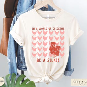 May include: A white t-shirt with the text "In a world of chickens, be a silkie" in red and pink lettering. The shirt features a red silkie chicken illustration surrounded by pink chickens.