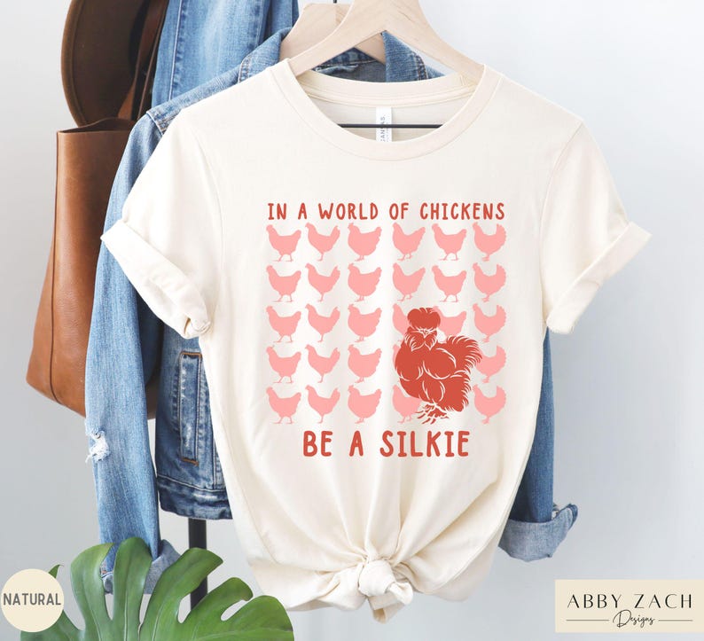 May include: A white t-shirt with the text "In a world of chickens, be a silkie" in red and pink lettering. The shirt features a red silkie chicken illustration surrounded by pink chickens.