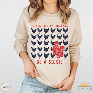 May include: A tan sweatshirt with the text "In a world of chickens be a silkie" in red and blue lettering. The design features a red silkie chicken surrounded by blue chickens.