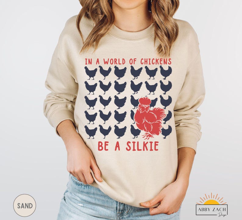 May include: A tan sweatshirt with the text "In a world of chickens be a silkie" in red and blue lettering. The design features a red silkie chicken surrounded by blue chickens.