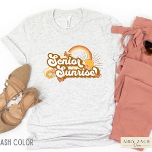 Puede incluir: Una camiseta gris con un dise&ntilde;o gr&aacute;fico retro que dice "Senior Sunrise" con un arco&iacute;ris, un sol y mariposas. El dise&ntilde;o est&aacute; en tonos naranja, amarillo y marr&oacute;n.