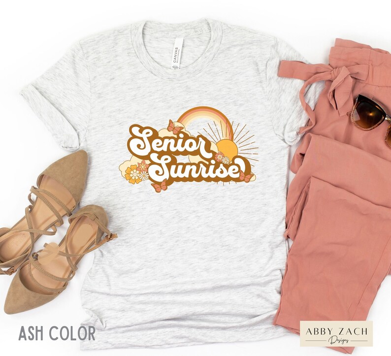 Puede incluir: Una camiseta gris con un dise&ntilde;o gr&aacute;fico retro que dice "Senior Sunrise" con un arco&iacute;ris, un sol y mariposas. El dise&ntilde;o est&aacute; en tonos naranja, amarillo y marr&oacute;n.