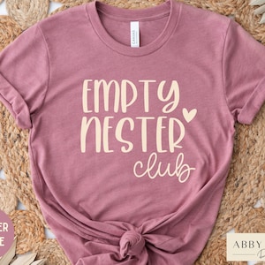 May include: A heather mauve t-shirt with the text "Empty Nester Club" in a cream color. The text is in a handwritten font with a small heart next to the word "Nester".