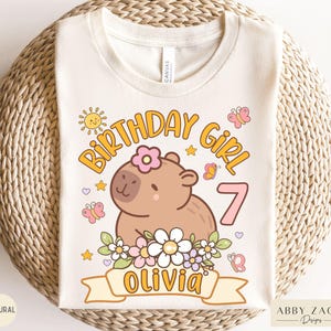 May include: A white t-shirt with a birthday girl design featuring a cartoon capybara with flowers, butterflies, and the text "Birthday Girl 7 Olivia".