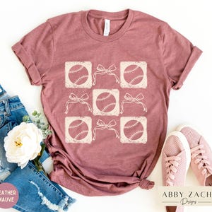 May include: A heather mauve t-shirt with a graphic design of baseballs and bows. The design is in a light cream color and is repeated across the shirt. The shirt is shown on a white background.