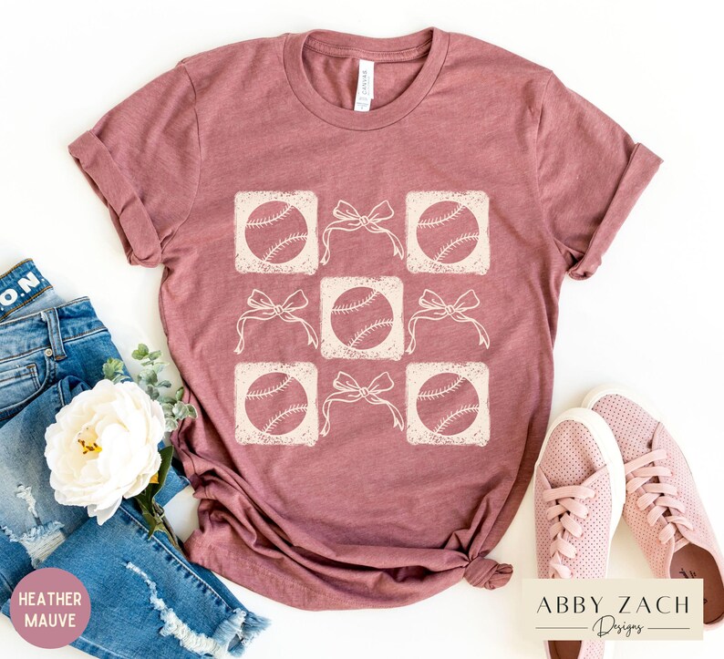 May include: A heather mauve t-shirt with a graphic design of baseballs and bows. The design is in a light cream color and is repeated across the shirt. The shirt is shown on a white background.