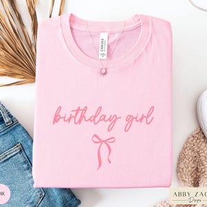May include: A light pink t-shirt with the words "birthday girl" in a cursive font and a bow graphic. A silver necklace with a sun pendant is on the shirt. The shirt is folded and placed on a white surface next to a pair of blue jeans.