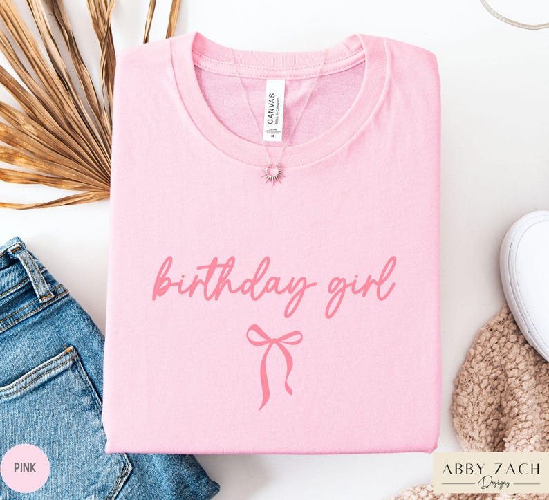 May include: A light pink t-shirt with the words "birthday girl" in a cursive font and a bow graphic. A silver necklace with a sun pendant is on the shirt. The shirt is folded and placed on a white surface next to a pair of blue jeans.