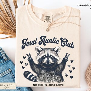 May include: Ivory-colored t-shirt with the text "Feral Auntie Club" and a raccoon graphic giving the peace sign. The shirt also has the text "NO RULES, JUST LOVE". A gold necklace is draped on the shirt.