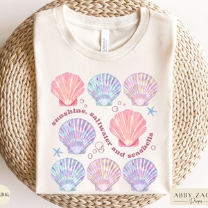 May include: Cream-colored t-shirt featuring a seashell design in pink, blue, and purple hues. The text "sunshine, saltwater and seashells" is printed in a curved font. The shirt has a crew neck and short sleeves.