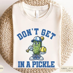 Puede incluir: Una camiseta blanca con un pepinillo de dibujos animados azul y verde que sostiene un bate de b&eacute;isbol y una pelota de b&eacute;isbol amarilla. El texto "Don't Get In A Pickle" est&aacute; escrito en letras azules alrededor del pepinillo.