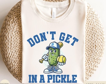 May include: A white t-shirt with a blue and green cartoon pickle holding a baseball bat and a yellow baseball. The text "Don't Get In A Pickle" is written in blue letters around the pickle.