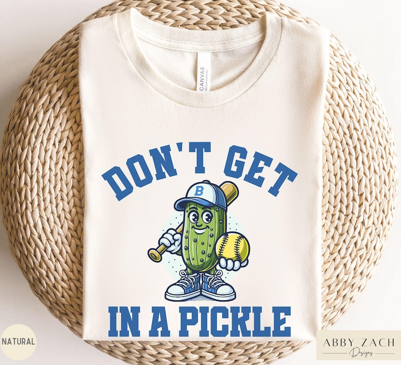 Puede incluir: Una camiseta blanca con un pepinillo de dibujos animados azul y verde que sostiene un bate de b&eacute;isbol y una pelota de b&eacute;isbol amarilla. El texto "Don't Get In A Pickle" est&aacute; escrito en letras azules alrededor del pepinillo.