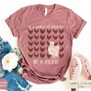 May include: A heather mauve t-shirt with the text "In a world of chickens, be a silkie" and a graphic of a silkie chicken surrounded by regular chickens.
