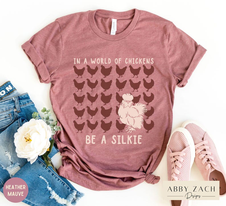 May include: A heather mauve t-shirt with the text "In a world of chickens, be a silkie" and a graphic of a silkie chicken surrounded by regular chickens.