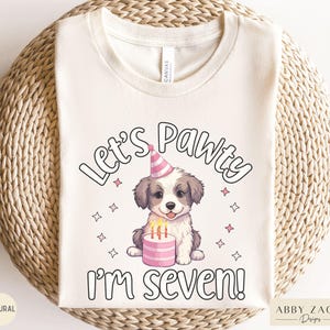 May include: A white t-shirt with the text "Let's Pawty I'm Seven!" in black. A cartoon dog wearing a pink party hat sits next to a pink cake with candles.